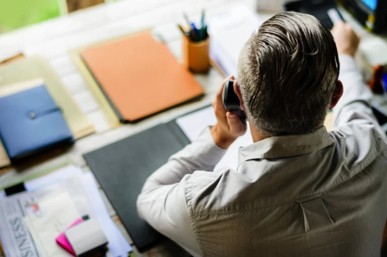 Imagem de um homem, visto de trás, a falar ao telefone, com trabalho organizado numa mesa no fundo